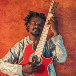 A Black man leans against an orange wall with his eyes closed. He is holding upright and strumming a red acoustic guitar with a cutout top that ends in a point in either side of the neck.