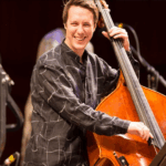 A laughing man with brown hair wearing a brown plaid shirt stands playing a standup bass