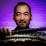 A close up of an Asian man with close trimmed hair and goatee against a glowing purple background