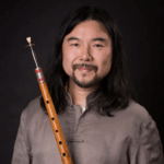 An Asian man with shoulder length black hair and a goatee smiles. He is holding a wooden flute upright against his right shoulder.