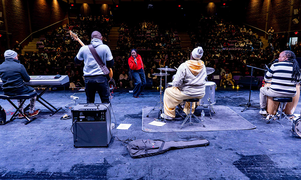 A Black woman with long braids and wearing a loose bright red sweater sings into a microphone while facing her band on a stage. Behind her is an auditorium full of students