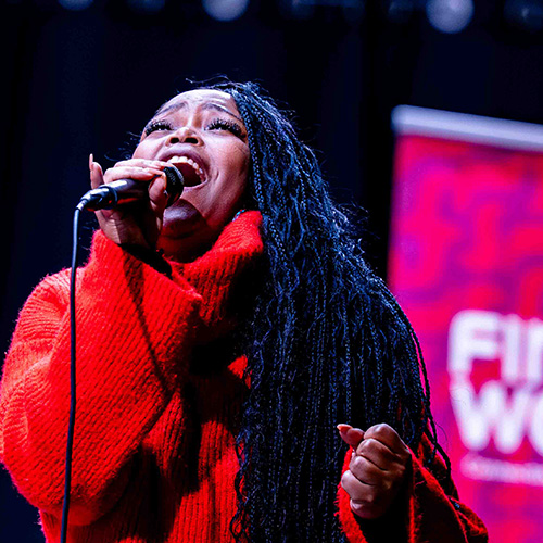 A Black woman with long braids and wearing a loose bright red sweater sings into a microphone with her face pointed up towards the ceiling. A pink and white FirstWorks logo banner is behind her