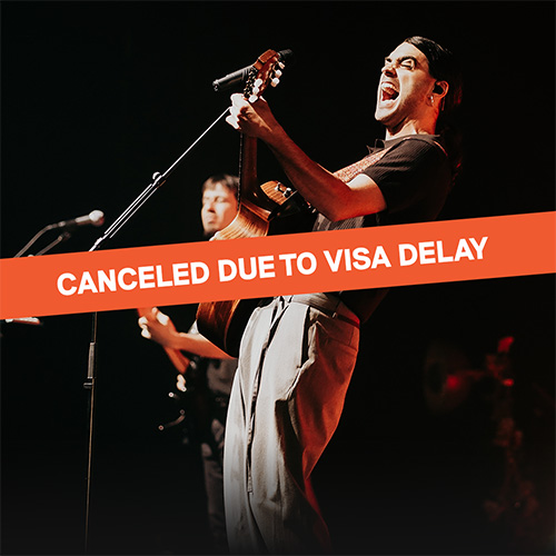 White text on orange banner reads CANCELED DUE TO VISA DELAY over a photograph of a young man playing guitar and singing passionately into a stand microphone on a stage. He is leaning back with his eyes closed and his mouth wide open. Another musician playing the bass is partially visible behind him.