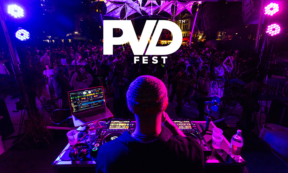 A DJ, shown from behind, spinning records for a large crowd of dancing people in the night time at the Providence Rink. The white PVDFest logo is overlaid on top of the photograph