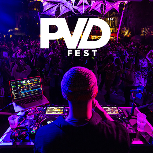 A DJ, shown from behind, spinning records for a large crowd of dancing people in the night time at the Providence Rink. The white PVDFest logo is overlaid on top of the photograph