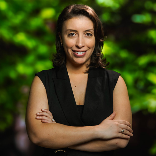 A smiling woman with dark brown hair and eyes and olive skin stands with her arms crossed over her chest in front of a blurred green leafy background. She is wearing a sleeveless black top.